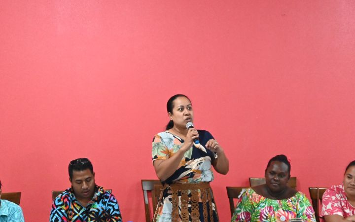 A group of men and women from the Pacific small island developing states are seated on a long table. On the left side are two men followed by a woman standing on the center holding a microphone as she speaks. On the right side just beside the standing woman are two women listening to the discussion. The background is a wall painted in red.