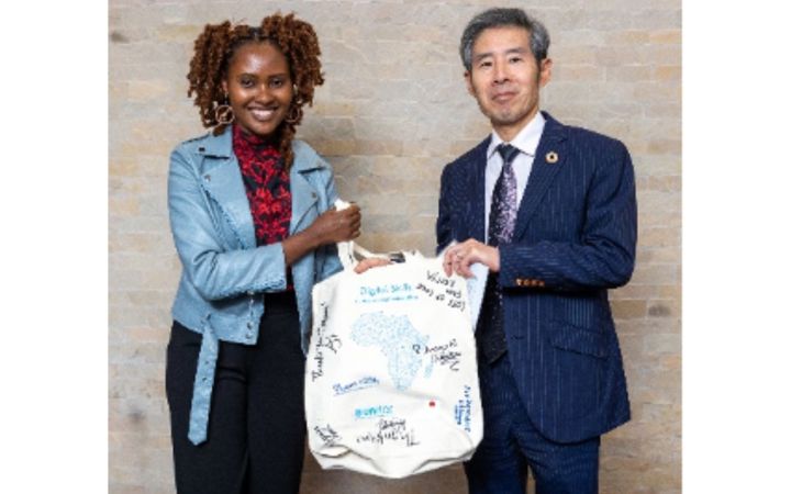 A young, dark-skinned woman with reddish brown braids atop her head and wearing a blue leather jacket stands with a grey-haired, angular East Asian man wearing a dark blue suit. The two hold up a large canvas bag printed with an African map and signed by people.