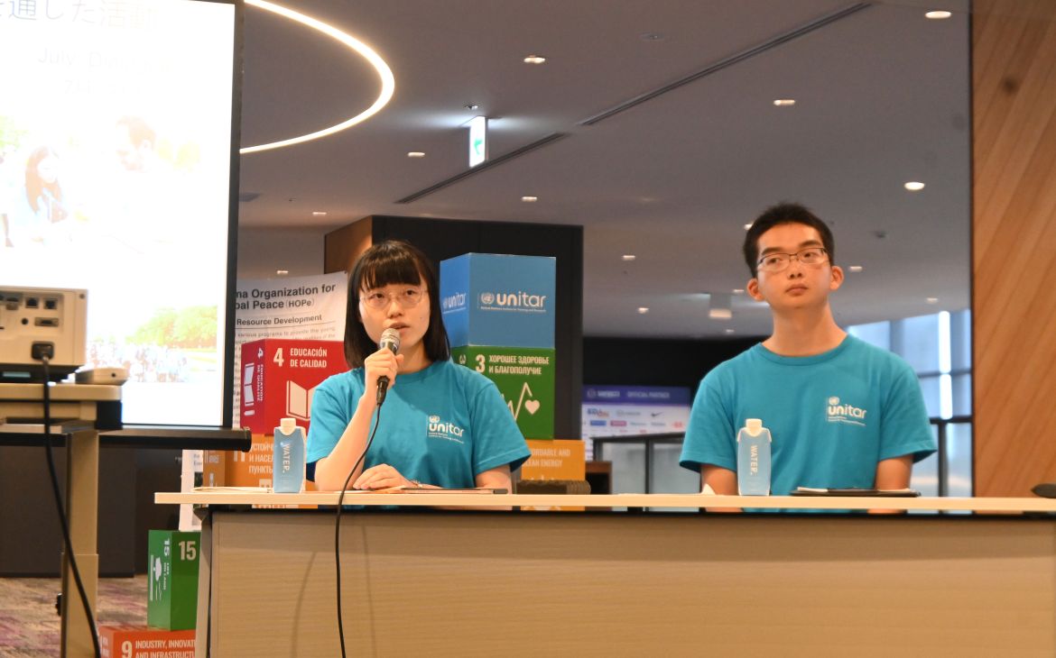 Two young particpants sitting behind a long brown table and one speaking into a microphone