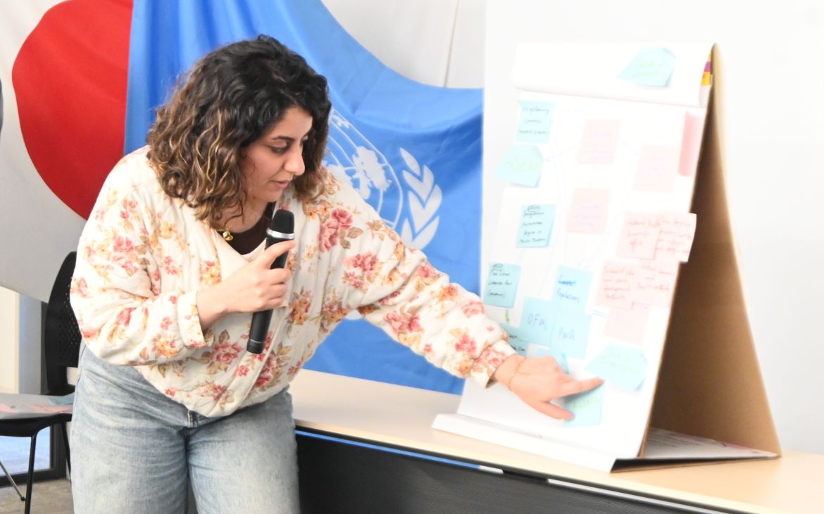A participant using a microphone and facing a panel and pointing to a handwritten memo on a paper panel