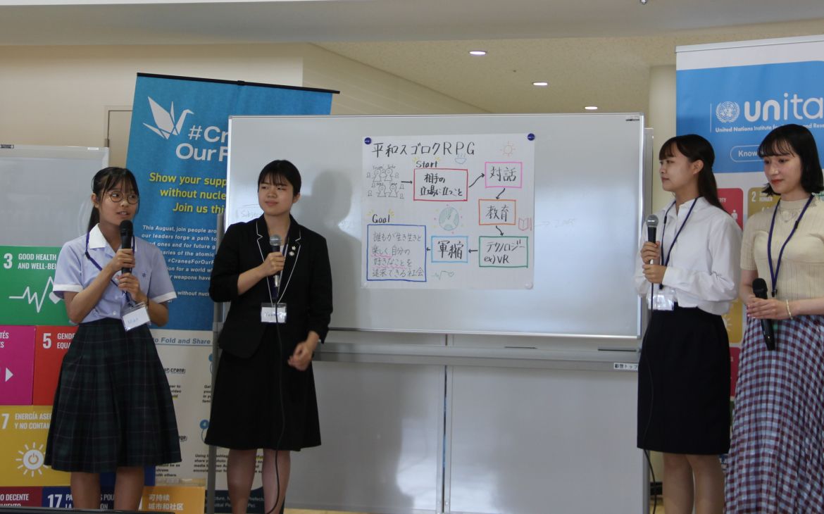 A photo of a group of four Japanese female students presenting in front of an audience (not shown). The student on the leftmost speaks while a fellow student on her left and the other two group members across them listens to her intently. The backgound is a whiteboard with a large white paper pasted on it containing some illustrations words written in Japanese while the left and right side of the background consists of the UNITAR logo and the SDG goals logos.