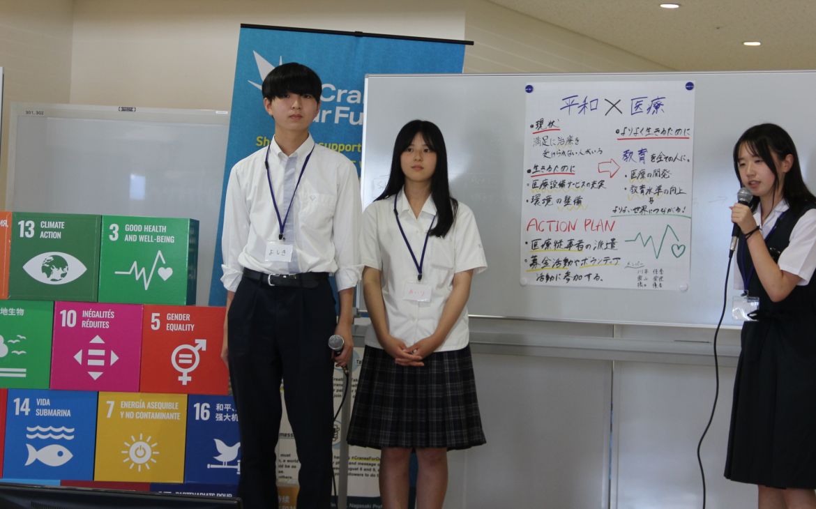 A group of Japanese students consisting of two female and one male presents in front of an audience (not shown). The female student on the right is speaking while the other members standing on the left side looks to the audience. The backgound is a whiteboard with a large white paper pasted on it containing some illustrations words written in Japanese with the SDG goals logos visible on the left side of the photo.