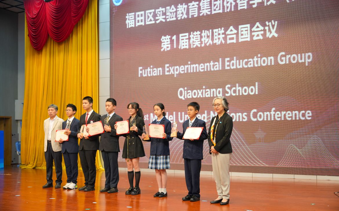 Six Chinese students in uniforms stand in a row on a big stage and hold up their certificates. An older man and woman stand next to the students.