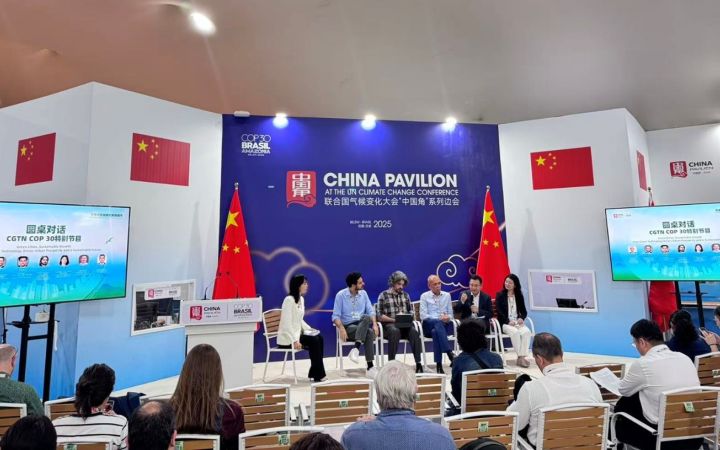 A wide-angle shot of the China Pavilion stage with two women and four men panellists seated casually on the stage. One is speaking.