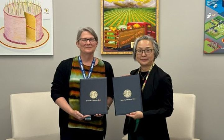 Two smiling professional women with greying hair, both hold up black, gold-embossed folders to the camera.
