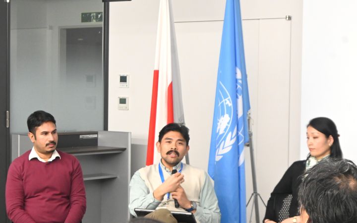 Three participants sitting in front of a crowd with a Japanese flag and UNITAR flag in the background and the person in the middle speaking through a microphone