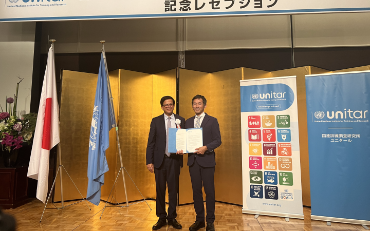 A photo of two men standing and wearing suits. The man on the left wearing eyeglasses poses beside the man holding a certificate. Behind them are the flags of Japan and the United Nations (left side of the photo) and two standees each featuring the SDGs and the logo of UNITAR.