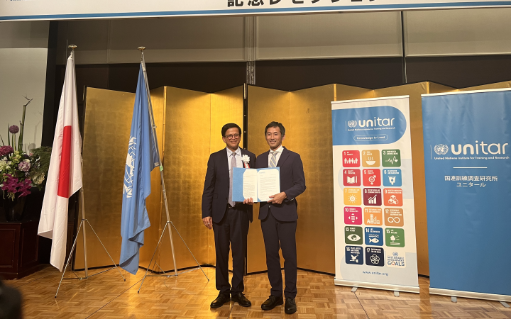 A photo of two men standing and wearing suits. The man on the left wearing eyeglasses poses with the man on the right who is holding a certificate. Behind them are the flags of Japan and the United Nations (left side of the photo) and two standees each featuring the SDGs and the logo of UNITAR. At the backmost is a stretch of gold-painted collapsible panels.