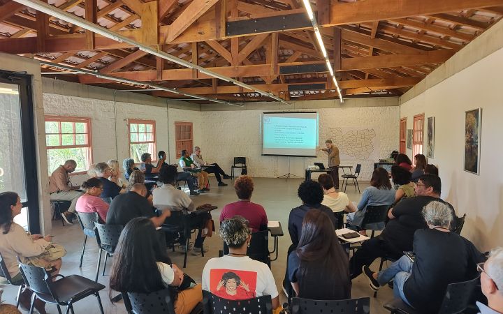 Participants of the Green Helmets training seated in a semicircle in a wooden-roofed room at UMAPAZ, listening to a speaker presenting slides, with a map of Sao Paulo’s East Zone displayed on the wall.