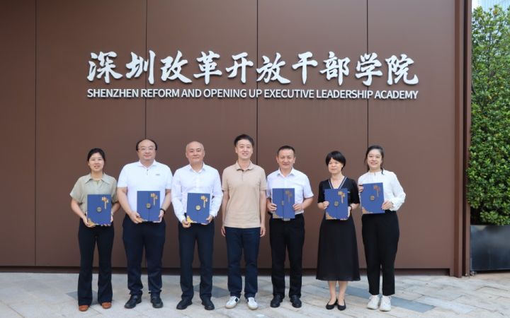 Seven Chinese men and women pose with blue folders in front of a sign for a Shenzhen executive leadership academy.