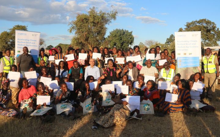 Three rows of smiling Zambian women and men pose in a grassy space, holding up their course certificates.