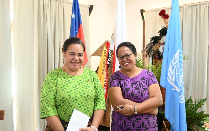 Two women from the Pacific smiles with the United Nations flag and what seems to be the flag of Samoa behind them hanging on flag standees. The woman in the left wears a green dress while the woman on the right is in purple.