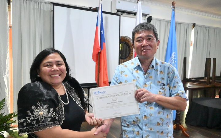 A Filipina wearing a traditional dress coloured in black and pearl necklace poses for a photo with a Japanese adult man as they hold a certificate.