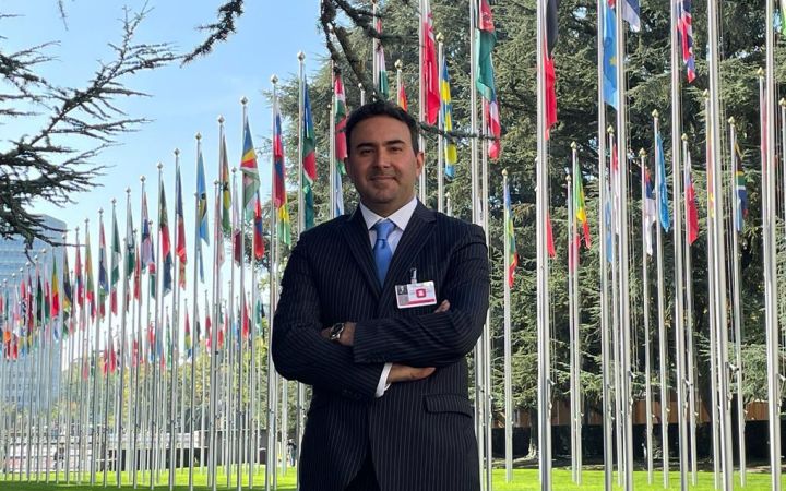 Dr. Sameh Al Muqdadi smiling with arms crossed, behind him are the flags of various countries at the UN Headquarters