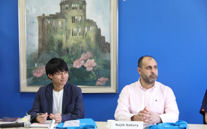 A photo featuring of two men looking to the right while listening to the meeting speaker (not visible on the photo) with their arms resting on a table with blue UNITAR bags, a microphone and a nameplate on a plastic standee. Their background is a blue wall with a hanging painting featuring the Hiroshima Bomb Dome and pink flowers.