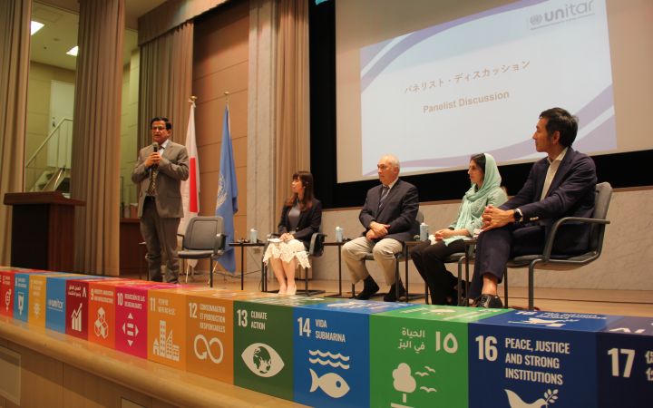 Close-up shot of the SDG goals placed at the stage floor while a group of four people comprised of two women and two men are sitting and looking to a man standing on the left side of the stage while holding a microphone and speaking.