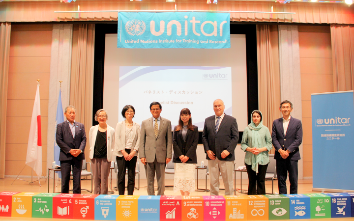 A group featuring men and women standing alternately on stage for a photo with a backdrop of UNITAR logo, the flags of Japanese and the United Nations. At the bottom of the stage are cube figures featuring the 17 SDGs.