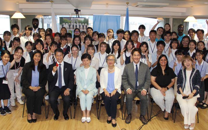 A group of Japanese people comprised of teenage students (back rows) and adult men and women in their corporate attires (sitting in front) poses for to have their photo taken. Among the crowds are men and women from different nationalities wearing blue UNITAR shirts.