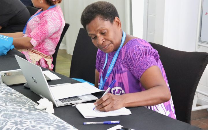Call for Applications: UNITAR Women's Leadership in Disaster Risk Reduction for World Tsunami Awareness Day 2026 A participant with a purple outfit sitting on a black chair, writing down notes and a laptop open next to the notebook