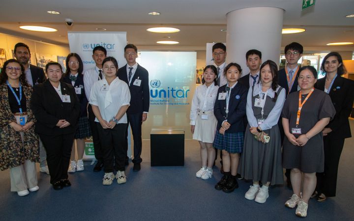 Group photo of 15 students and professionals, standing in two rows around a glass UNITAR sign in a building lobby