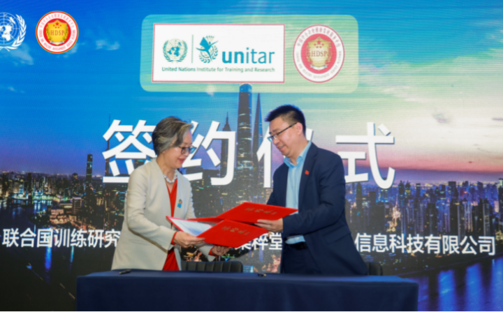 An Asian woman and an Asian man in suits exchange two red folders in front of a backdrop with UNITAR and JICUITANG logos and the words “signing ceremony” printed in Chinese on a photo of Shanghai at sunset.