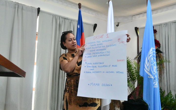 A woman from a Pacific small island developing state stands in front an audience (not shown) as she holds a large piece of white paper containing some texts which she is reading. Behind her are the flags of Samoa and Japan (both partially hidden) and the United Nations flag – all hanging on flagpoles.