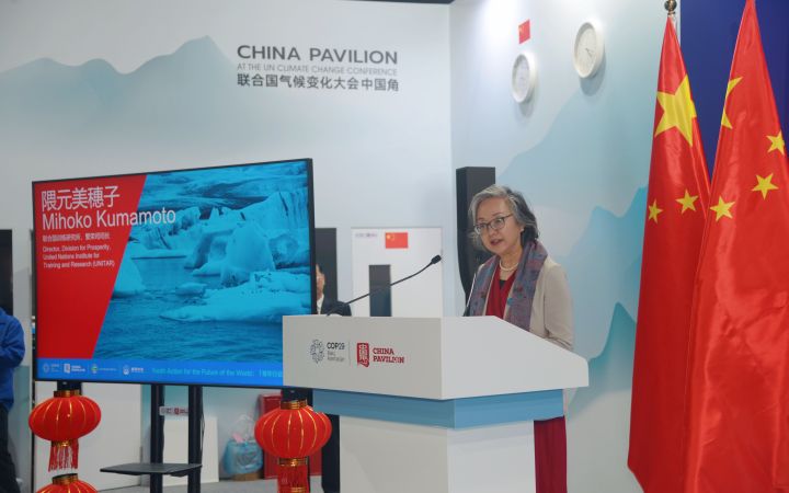 Asian professional woman with silver hair speaking at a podium in front of a display with two Chinese flags, a flat screen, and red Chinese lanterns.