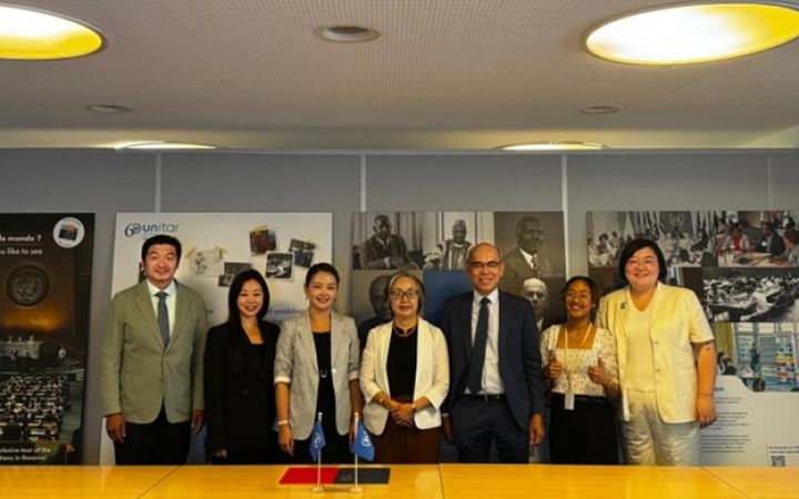 Five business women and two men stand smiling at a table with small UN flags, in front of a wall of photo panels about UNITAR.