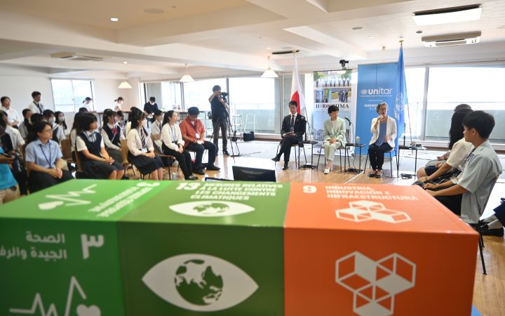 A photo featuring three Japanese adults (from left to right: a man wearing black suit, a woman in a mint green corporate dress, and a woman wearing white blazers with blue inner blouse). On the leftmost and rightmost sides of the photo are students while the foreground shows the SDGs 3, 13 and 9 cubes (zoomed. The background behind the panellists includes the Japanese flag, a banner of Hiroshima Prefectural Government, UNITAR logo, and the UN flag.