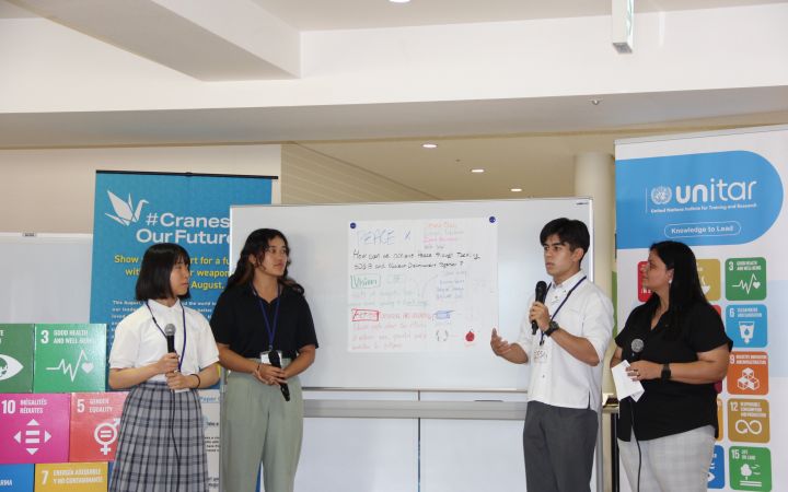 A photo of a group of Japanese students presenting in front of an audience (not shown). On the left side are two female students holding microphones as they listens to the male student speaking on the right. Right beside the male student is what seems to be a event co-facilitator. The backgound is a whiteboard with a large white paper pasted on it containing the keywords from the presenters while the left and right side of the background consists of the UNITAR logo and the SDG goals logos.