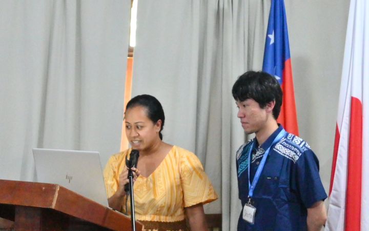A photo of a woman wearing a yellow dress and a man in blue short sleeve polo standing in front of an audience (not shown). The are standing beside a rostrum (left) where a laptop is placed. The woman speaks using a microphone while reading something on the laptop. The man beside her (right) is looking on the laptop screen as well. Behind themm are the flags of Samoa and Japan (partially hidden) hanging on flag stands.