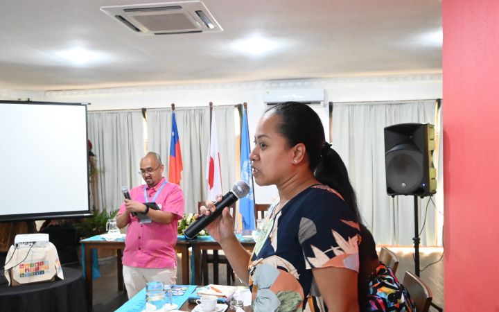 A woman from a Pacific small island developing state stands in front of a group of people (not shown) while holding a microphone as she speaks. Behind her is a man wearing a pink polo shirt and the Samoa, United Nations and Japan flags hanging on flag poles.