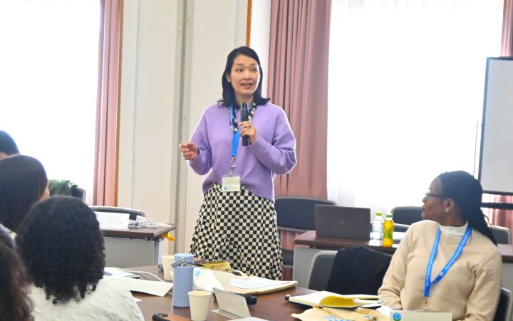 A participant standing up in front of her group session table to speak through a microphone and other participants sitting and looking at her
