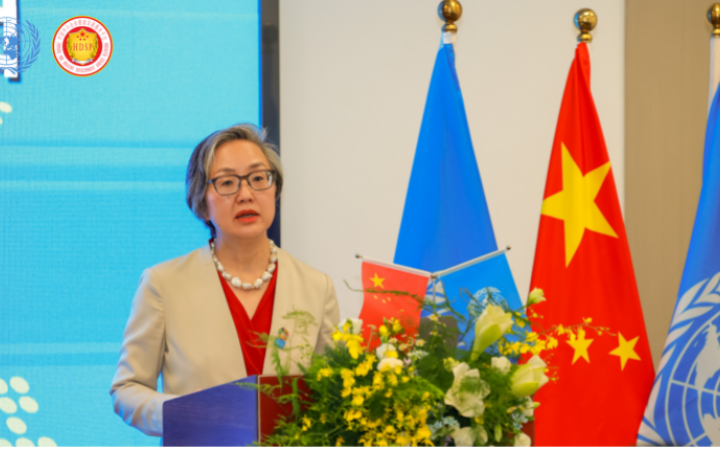 Asian woman wearing glasses and a beige suit over a red shirt speaks at a podium decorated with big flowers and a small UN flag and Chinese flag. Behind her stand big flags of the United Nations and China.