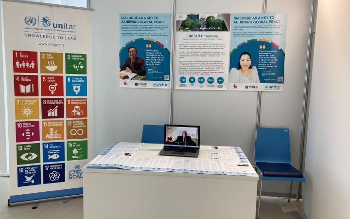 An exhibit space featuring a vertical banner on the left with the SDGs. There are three smaller posters on the wall (center) featuring a male, an information about UNITAR's training programme, and a woman. On the lower center is a white table where a laptop is placed in the center surrounded by flyers.