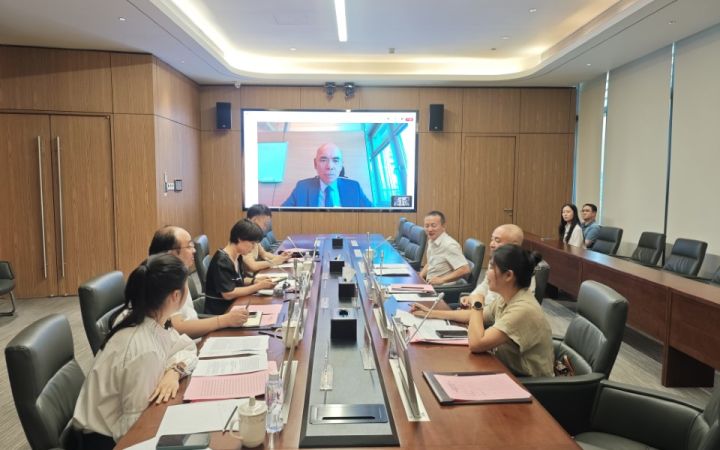 East Asian professional women and men sit around a long conference table, with a screen on the wall showing a man on a video call