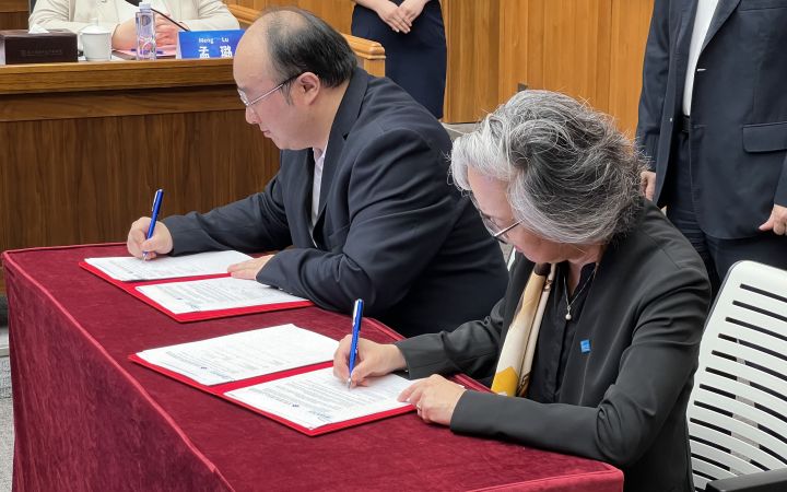 A man and grey-haired woman sign documents at a red-velvet-covered table as others look on.