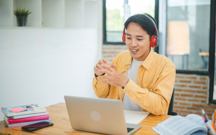 A stock photo image of a smiling person with a red headphone gesturing to a laptop computer 